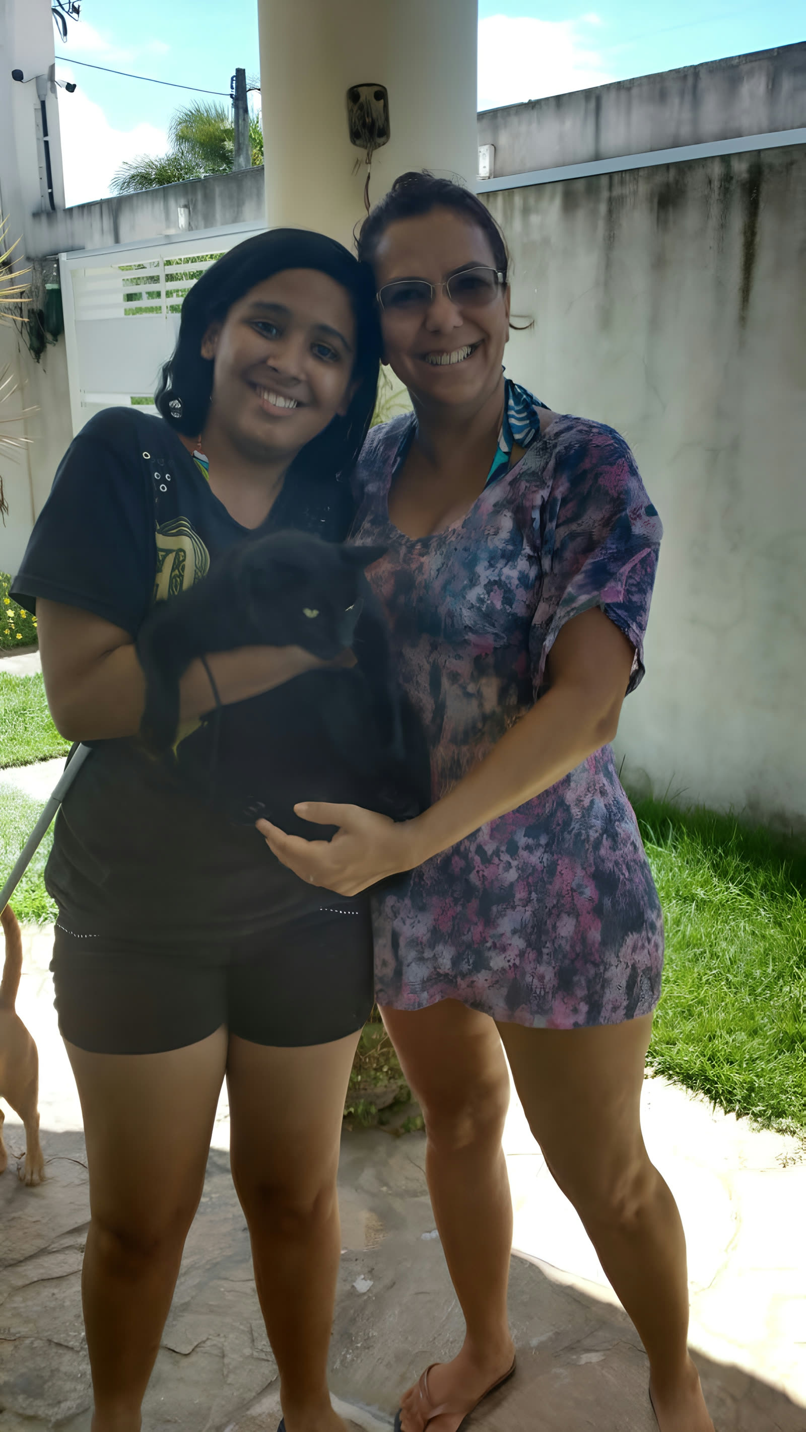 Louise, com um gato preto no colo, abraçada com Vânia em um quintal ensolarado.